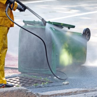 Sanitation worker uses a power washer to clean a compost recycling green bin.